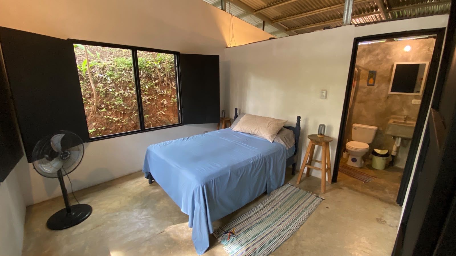 Bedroom with a double bed, standing fan and shuttered window, with an ensuite bathroom through an open door