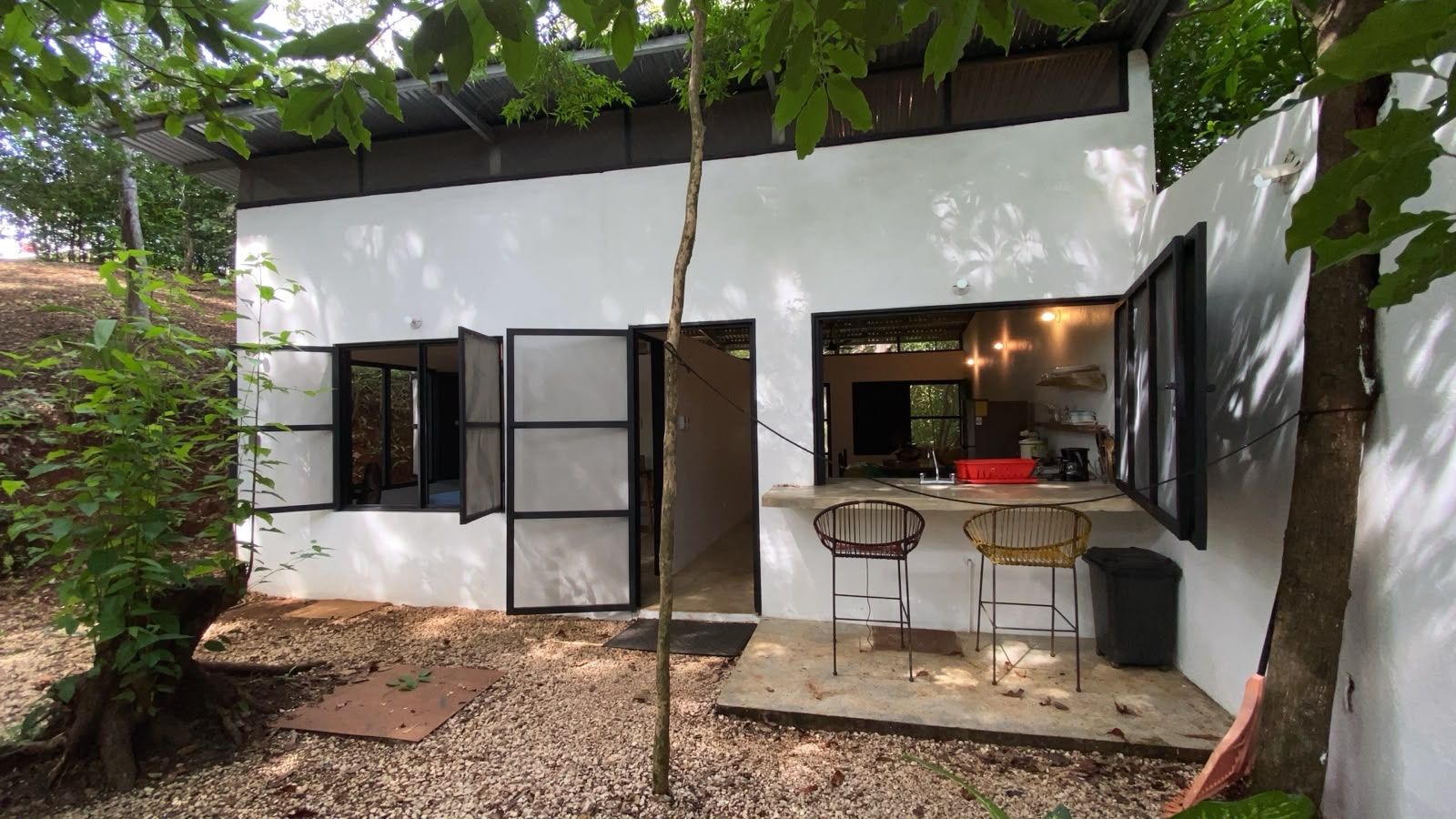 White single-storey house among trees with open windows and an outdoor counter with two bar stools by the kitchen