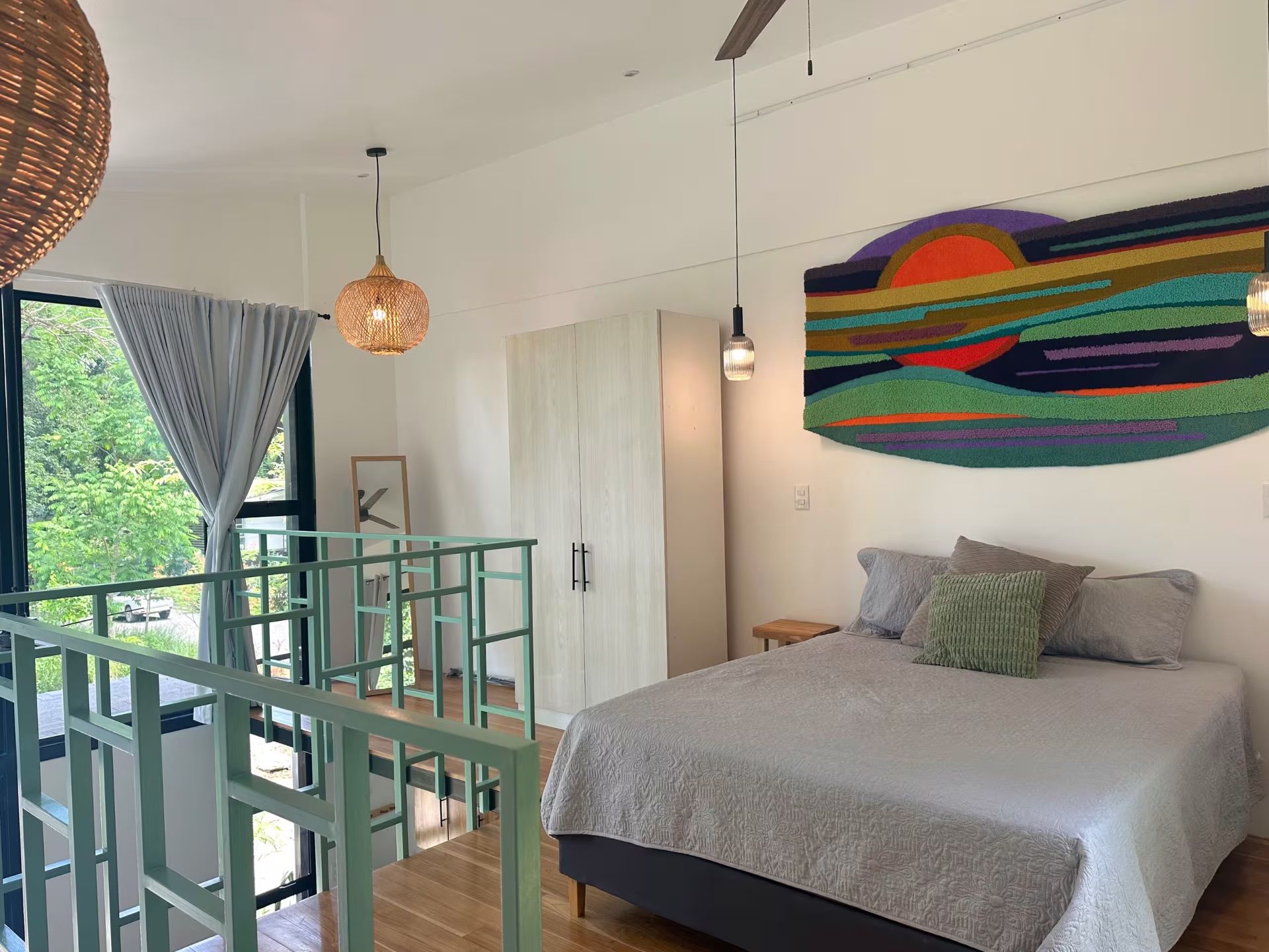 Loft bedroom with a queen bed, wardrobe and ceiling fan, overlooking the stairs and a window to trees