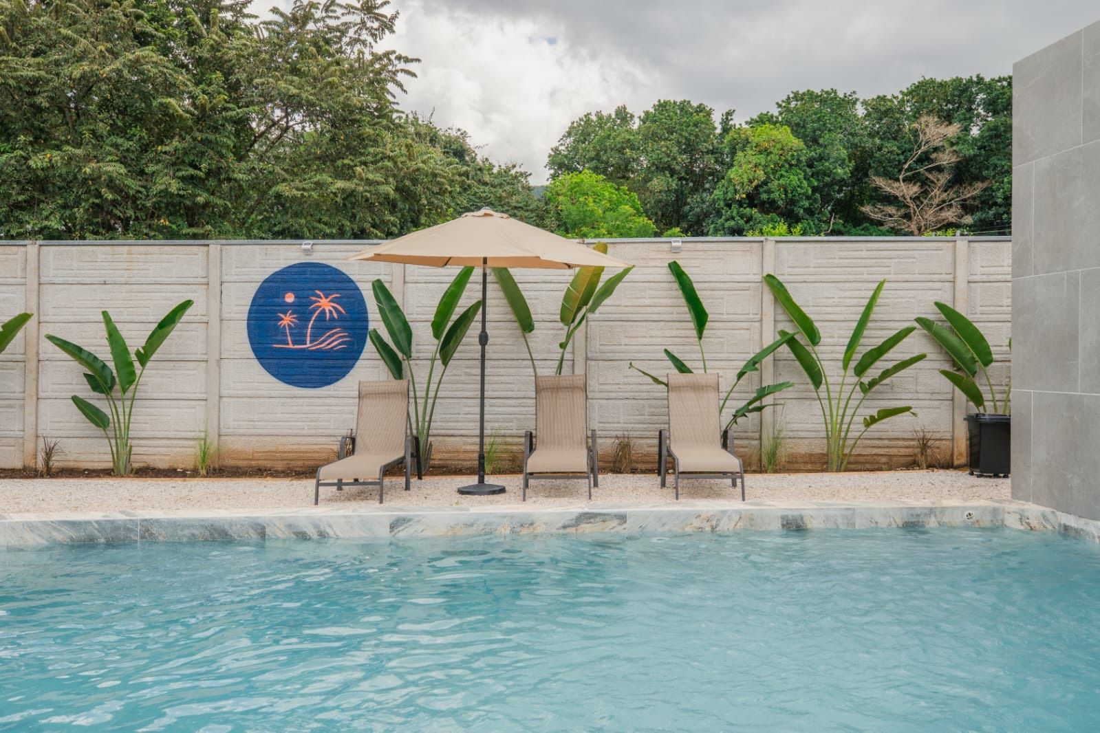 Fenced pool with three lounge chairs and a sun umbrella against a wall lined with banana plants