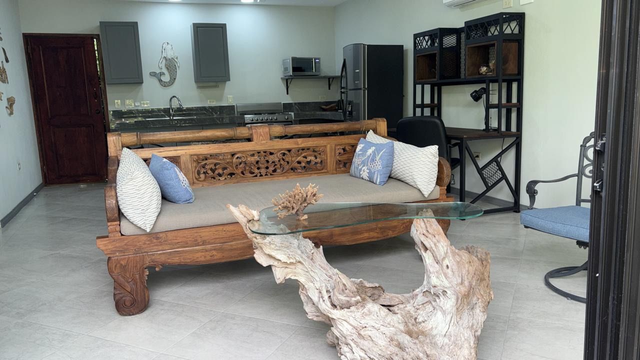 Open living space with a carved wooden sofa and driftwood coffee table, kitchen and desk behind