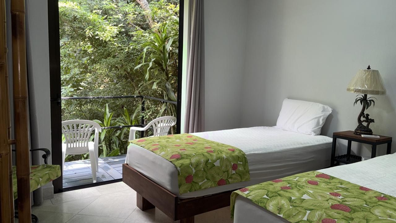 Bedroom with two single beds and a glass door opening onto a balcony with two chairs and jungle beyond