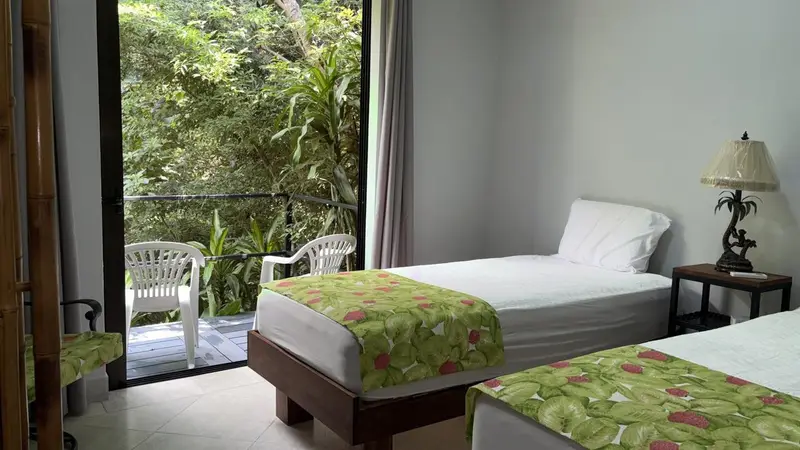 Bedroom with two single beds and a glass door opening onto a balcony with two chairs and jungle beyond