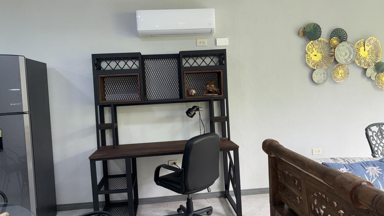 Desk with shelving, office chair and lamp beneath a wall-mounted A/C unit, next to the fridge